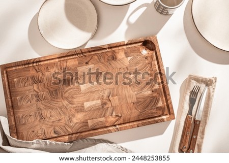 A wooden cutting board next to ceramic plates and silverware wrapped in a linen napkin, bathed in sunlight.