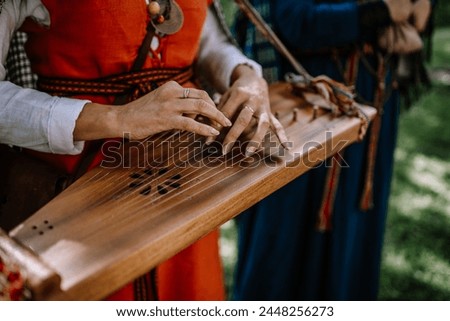 Valmiera, Latvia - July 14, 2023 - Hands playing a kokle, a traditional Latvian stringed musical instrument, with musicians in folk costumes blurred in the background. Royalty-Free Stock Photo #2448256273