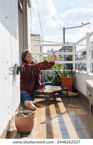 Latin adult woman on terrace of her apartment taking selfie photo with her phone while sitting resting in red sweater, calmly reading book and drinking coffee, lifestyle concept