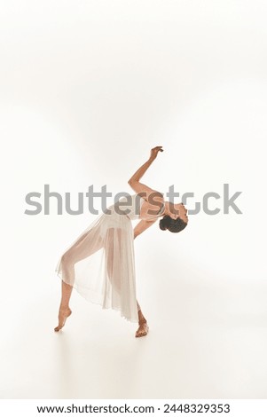 A graceful young woman in a flowing white dress performs in a studio setting against a white background.
