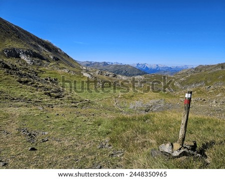 Photo of a hiking trail with a path in the mountain and a white and red marker post. Muntains at background and blue sky Royalty-Free Stock Photo #2448350965