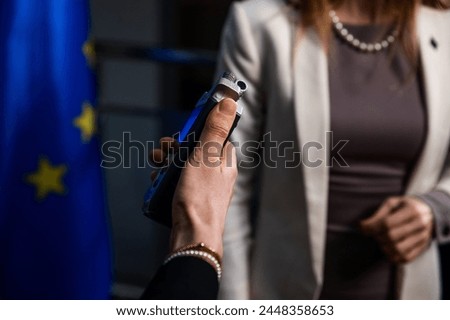 Close-up view of a voice recorder in the hand of a journalist which is aimed at a female politician with an EU flag Royalty-Free Stock Photo #2448358653