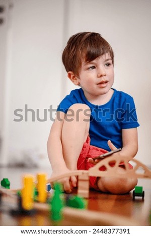 A joyful young boy with a smartphone sits amidst a wooden train setup, his bright laughter echoing the playful fusion of digital and tangible play Royalty-Free Stock Photo #2448377391