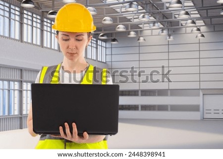 A female engineer is looking at a laptop. There is a production room in the background. The engineer looks at the work plan on a laptop.