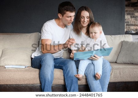 Family: mom, dad sitting on sofa in living room, reading book to baby, showing educational pictures. Young parents spend time with their baby, learning and communicating. Happy family concept, weekend