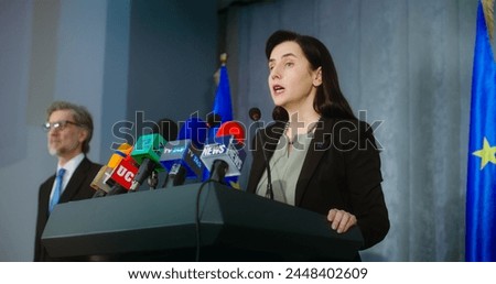 Female representative of the European Union during press conference. Confident politician makes an announcement, answers journalists questions and gives interview for media. Backdrop with EU flags. Royalty-Free Stock Photo #2448402609