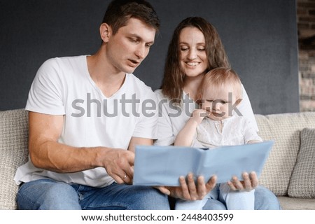 Mom, dad and child are sitting on sofa at home, reading together, looking at funny educational pictures in book. Family enjoying weekend at home. Concept of communication, learning, relaxation.