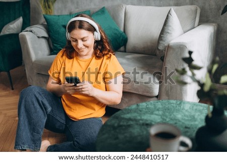 Smiling girl relaxing at home, she is playing music using a smartphone and wearing white headphones. Young woman hearing enjoying good audio sound lounge on couch at home.