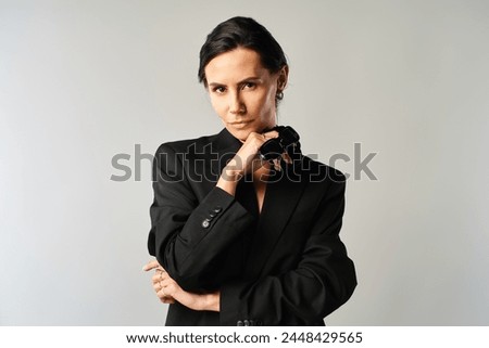 A fashionable woman in sunglasses confidently poses in a sleek black suit against a grey studio backdrop.