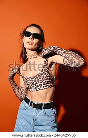 A fashionable woman in a leopard print top and sunglasses poses confidently in a studio setting against an orange background.