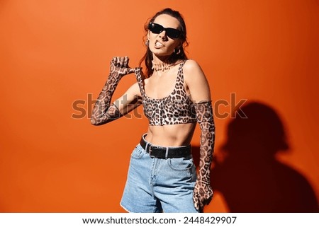 A stylish woman in sunglasses stands confidently in a studio, wearing a leopard print top and denim shorts against an orange background.