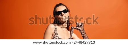 A stylish woman in a leopard print dress poses with a leopard print in a studio setting against an orange background.