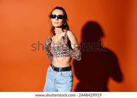 A stylish woman in sunglasses poses in a leopard print top and denim shorts against an orange background, exuding summertime fashion vibes.