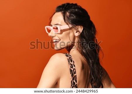 A fashionable woman exudes summertime vibes in a leopard print top, accessorized with pink sunglasses, against an orange background.