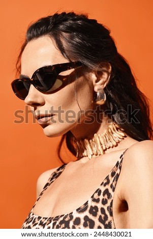 A stylish woman with sunglasses poses in a leopard print top against an orange studio background, exuding summertime fashion vibes.