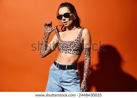 Stylish woman in sunglasses posing confidently in a leopard print top and jeans against an orange background.