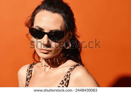 A fashionable woman wearing sunglasses and a leopard print top poses confidently in a studio against an orange background.