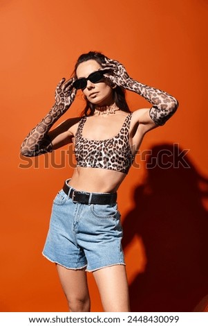 A fashionable woman in a leopard print top and denim shorts exudes confidence in a studio setting against an orange background.