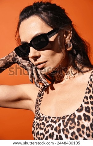 A stylish woman in a leopard print top and sunglasses poses confidently in a studio against an orange background.