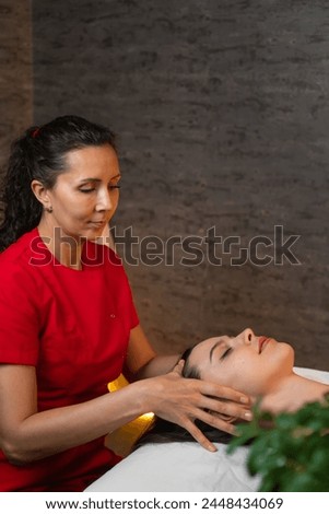 Masseur in a red uniform suit gives a head massage to a young brunette woman lying on a massage table in the spa solon. Spa concept, self-care. Vertical photo