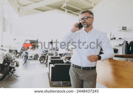 Male buyer man wears shirt prop up chin look camera standing at office table in dealership store indoors want to buy new motorbike or car talk speak on mobile cell phone. Business lifestyle concept Royalty-Free Stock Photo #2448437245