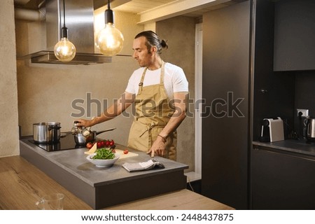 Handsome young man 40-44 years old, cooking dinner in modern kitchen interior. attractive guy, chef in beige apron, standing at kitchen counter with fresh organic ingredients, preparing delicious meal Royalty-Free Stock Photo #2448437875