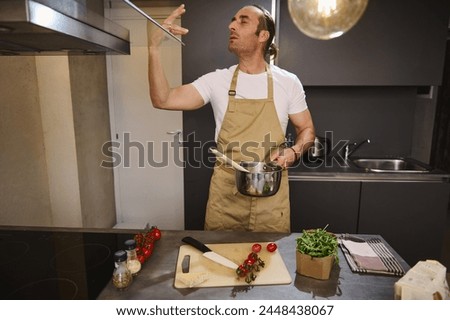 Handsome male chef holding saucepan with tomato sauce, kissing fingers, showing tasty hands sign, saying bellissimo cooking gourmet food in the home kitchen. People. Culinary. Alimentation. Lifestyle. Royalty-Free Stock Photo #2448438067