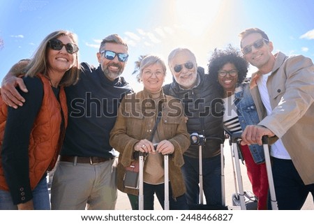 Portrait group of middle-aged tourist friends posing embraced smiling and looking at camera enjoy together journeys. Diverse people of tourism with luggage in European street on sunny day. Low angle