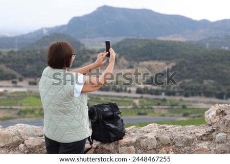 Rear view of mature woman traveler with backpack relaxes on hill, takes pictures of the mountains and valley landscape with smartphone. Concept of active lifestyle, hiking, trekking at summer holiday.