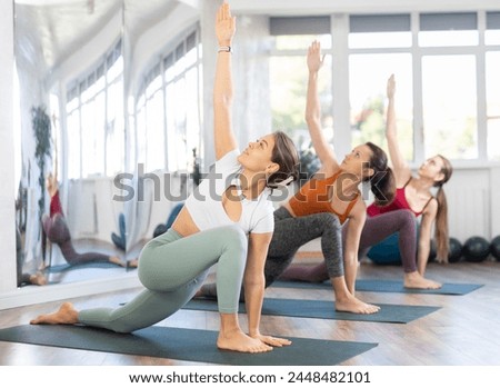 Positive young girl with group of female yoga enthusiasts performing Anjaneyasana with twist, Low Lunge with back knee grounded and torso twist, enhancing spinal mobility and core strength in studio