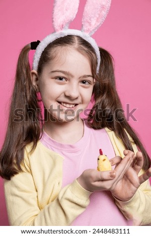 Young cute kid holding a stuffed chick in front of camera, positive excited girl feeling happy about easter celebrations. Small child with fluffy bunny ears smiling over pink background.
