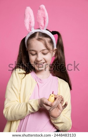 Small young kid showing a cute stuffed chick toy in studio, joyful little girl with golden arrangement over pink background. Youngster with sweet fluffy ears for easter festivity in front of camera.