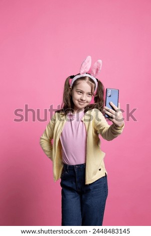 Positive cute schoolgirl taking pictures with her mobile phone in studio, smiling at camera and posing with confidence against pink background. Cheerful young kid takes photos for easter festivity.