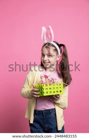 Little joyful child holding basket with painted eggs and a pink rabbit, celebrating easter festivity over pink background. Small cheerful kid with bunny ears showing spring celebration decorations.