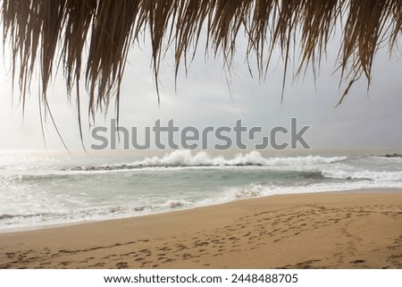 A tranquil sandy beach with a charming straw-roofed beach hut nestled under moody, cloud-filled skies. The crashing waves create a dramatic contrast as footprints dot the serene shoreline. Royalty-Free Stock Photo #2448488705