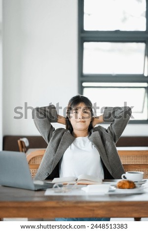 Relaxed freelancer leans back in her chair, taking a well-deserved break from work, with her laptop and notes laid out on the cafe table. Royalty-Free Stock Photo #2448513383