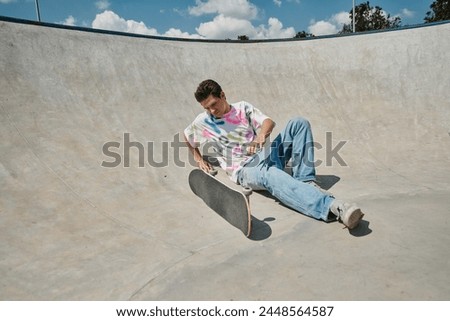 A young skater boy defies gravity, sitting near skateboard on ramp in a outdoor skate park on a sunny summer day.