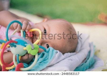 Infant Baby Girl is laying on her back in a pink bodysuit biting a development toy for teething.