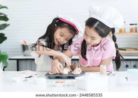 Two cute sisters with chef hat enjoy baking, decorating cupcake with jelly and icing sugar at kitchen at home. Little children girls have fun together. Happy Asian family