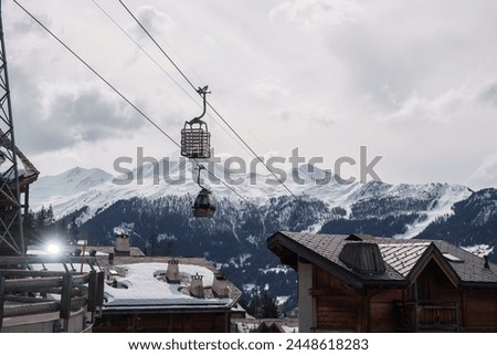 Winter wonderland scene at a ski resort with wooden chalet buildings, ski lift, snow covered mountains, and ski runs. Serene and chilly atmosphere. Located in European Alps or similar region. Royalty-Free Stock Photo #2448618283
