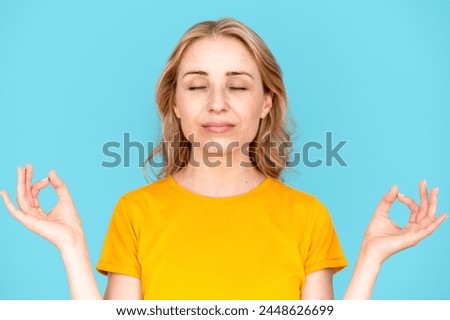 Satisfied woman keeping eyes closed and doing meditation gesture with fingers isolated over blue studio background. Trying to calm down. Relax and yoga concept.