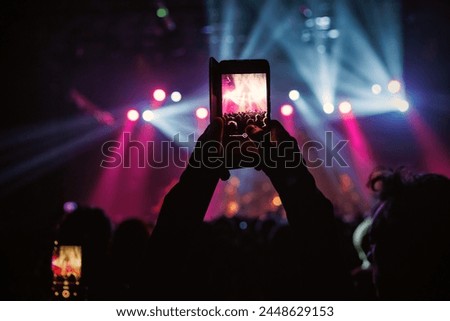 A hand holds a smartphone, recording a lively event illuminated by colorful lights.