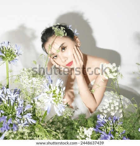 "The beautiful Asian bride proudly lifts her chin in front of a table of purple flowers, against a white backdrop for a studio photoshoot."