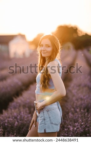 A young and slim girl walks and enjoying fun the time. Smiling female walking among lavender flowers with sunlight on summer day. Woman standing in lavender field and sniffing flowers at sunset.