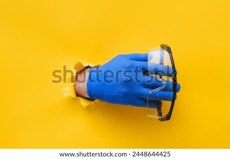 The male hand in a blue fabric work glove holds safety glasses. Torn hole in yellow paper. Good job, eye protection and safety concept. Copy space.