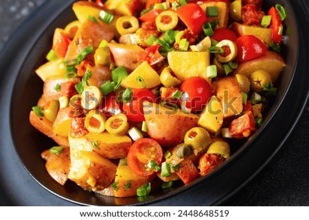 spanish potato salad of chorizo, green olives, cherry tomatoes, red pepper and cilantro with honey mustard dressing in  black bowl on concrete table, close-up, dutch angle view Royalty-Free Stock Photo #2448648519