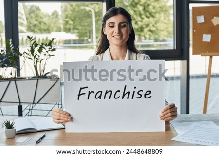 A businesswoman in a modern office sitting at a table and holding a sign with franchise letters
