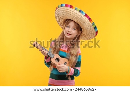 Cute girl in Mexican sombrero hat playing ukulele on orange background