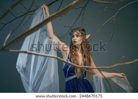 A young woman in a blue dress gracefully holds a delicate white veil in a magical studio setting fit for an elf princess. Royalty-Free Stock Photo #2448670175