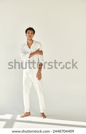 elegant african american man in white attire posing barefoot and looking at camera on beige backdrop